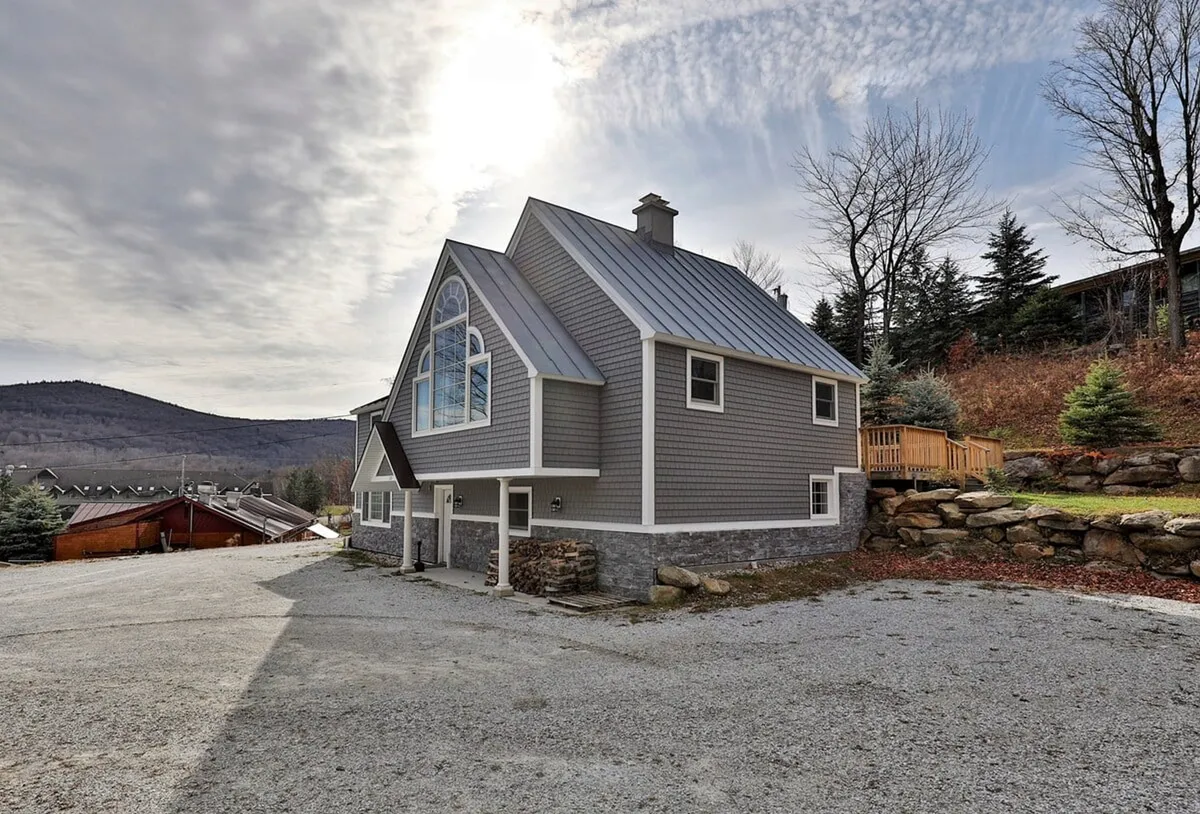 A wide exterior view focusing on the chalet’s large window with architectural details, gravel driveway, and stone foundation. The setting is serene, hinting at both accessibility and the tranquil atmosphere awaiting guests.