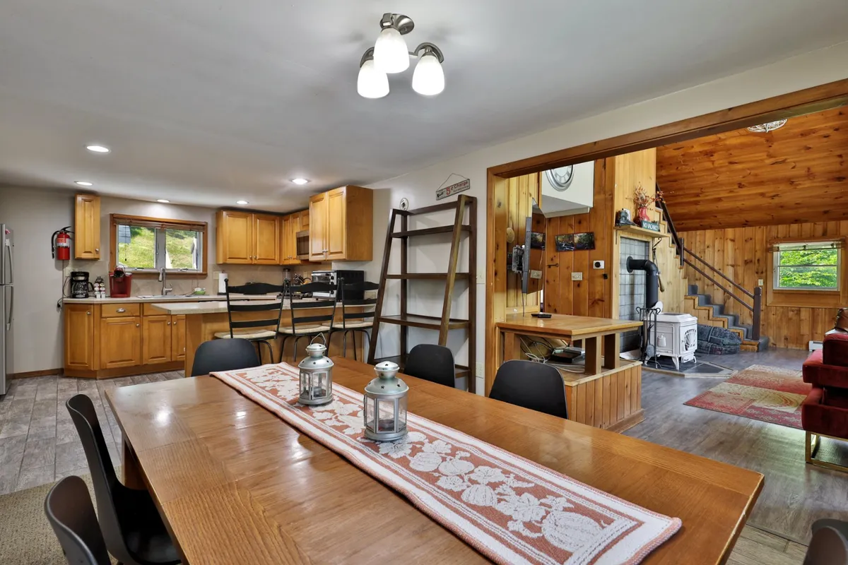 The image captures the adjacent dining area with a large wooden table covered by a decorative runner, surrounded by chairs. Wooden cabinets and a visible connection to the kitchen set the stage for shared meals, laughter-filled dinners, and game nights.