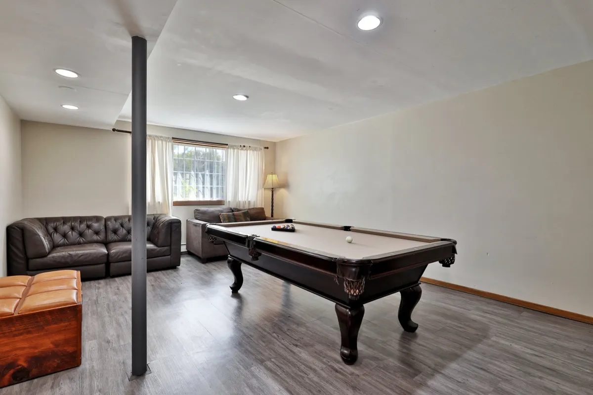 A friendly and relaxed game room featuring a pool table, chocolate brown leather couch, wooden ottoman, and a large window with curtains. The space encourages friendly competition, socialization, and celebration.
