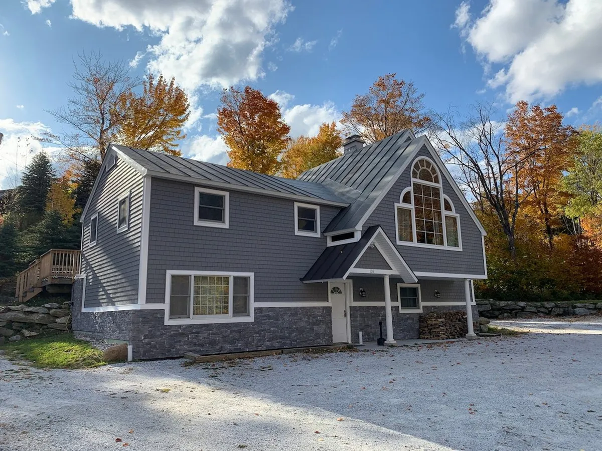 The image showcases the inviting facade of the West Park Chalet, framed by towering Vermont trees. Large windows reflect the natural surroundings, and a welcoming porch hints at spots perfect for sunset watching and gathering with friends or family. The setting radiates warmth, nestled in a peaceful woodland environment ideal for relaxation and adventure.