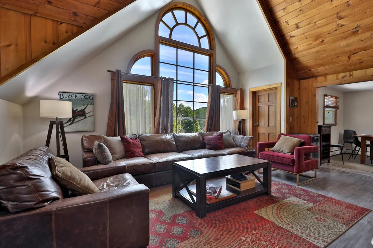 This image features a cozy sectional sofa arranged near a large arched window, with colorful accent chairs and a rustic wooden ceiling. The space is designed for comfortable gatherings, offering abundant natural light and a sense of warmth.