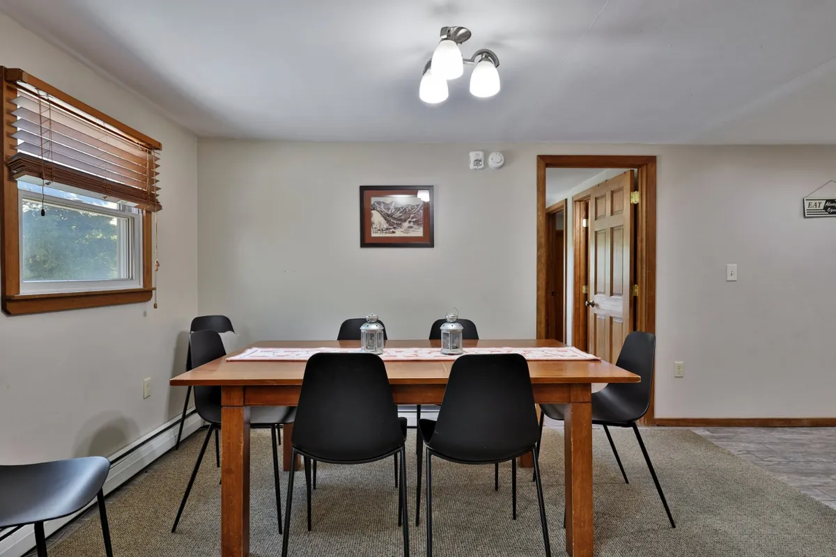 A dining area featuring a large wooden table surrounded by black chairs and wall art, positioned by a window. The table is set up for meals, game nights, or intimate group discussions, with natural light enhancing the welcoming environment.
