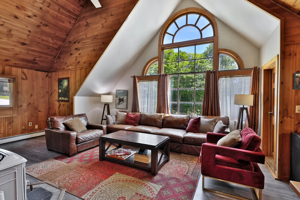 A cozy living area with an arched large window, a leather sectional sofa, a coffee table filled with books, red accent chairs, and wooden wall paneling. The arrangement encourages conversation and relaxation, with natural light streaming in, highlighting the welcoming atmosphere.