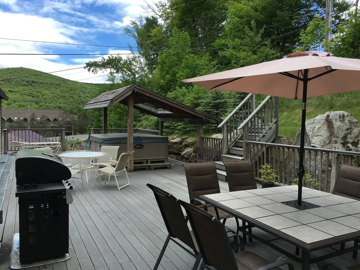 The image captures the spacious wooden deck featuring a dining table with chairs, a grill, a hot tub, and a large umbrella. The setting is designed for social gatherings, barbecuing, and relaxing outdoors, with the hot tub positioned for easy access from the deck.