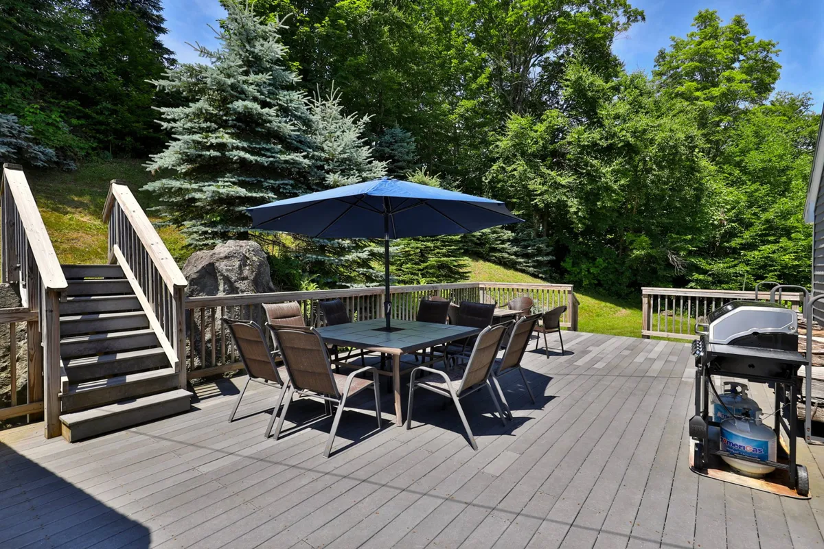 This image features an outdoor dining table with an umbrella, grill, nearby staircase, and trees encircling the area. It evokes scenes of casual barbecue dinners, group celebrations, and relaxed socializing under the open sky.