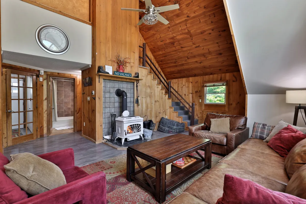 A welcoming living area with wooden walls, a fireplace, a wooden staircase, a coffee table, and sofas. The setting is ideal for group gatherings, sharing meals, and relaxing after a day of celebration.