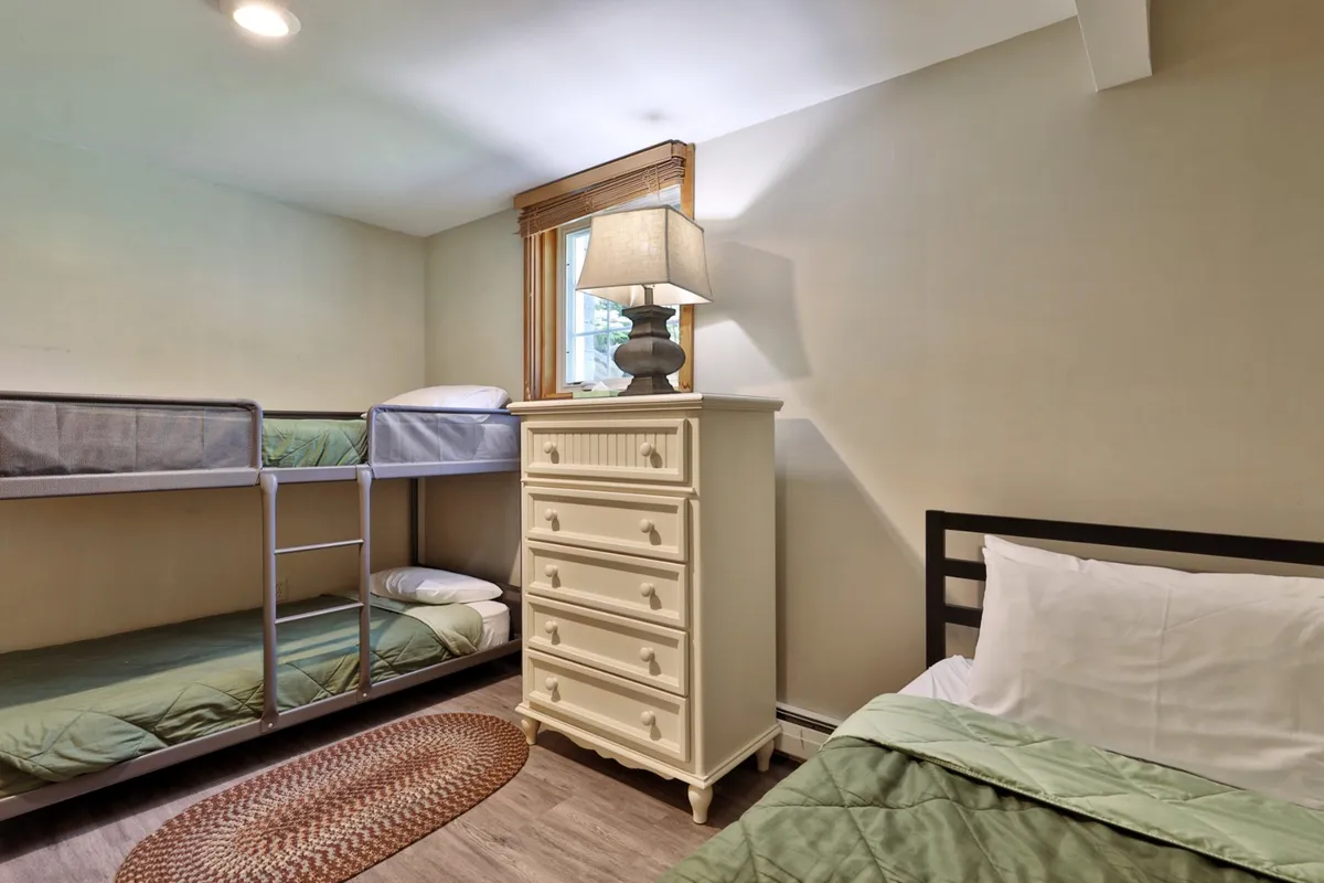 A bright and inviting bedroom featuring bunk beds, dressers, a lamp, and a bed with crisp white linens. Sunlight filters through the windows, creating a cheerful ambiance for families or children to enjoy sleepovers together.