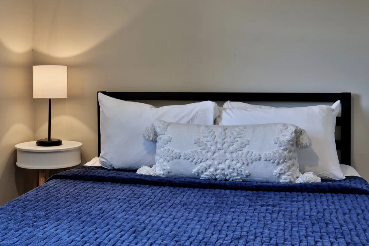 A serene bedroom with a comfortable bed, decorative pillow, and a bedside lamp. The lighting is soft, creating a relaxing atmosphere ideal for rest and reading. The space highlights warm blankets and a peaceful setting.