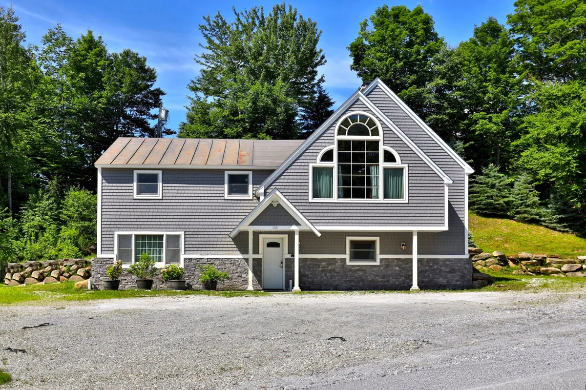 The image showcases the chalet's welcoming exterior, with a large front window, stone facade, and lush greenery surrounding the house. The scene captures the inviting atmosphere of the property at sunset, hinting at lively gatherings and relaxed outdoor moments.