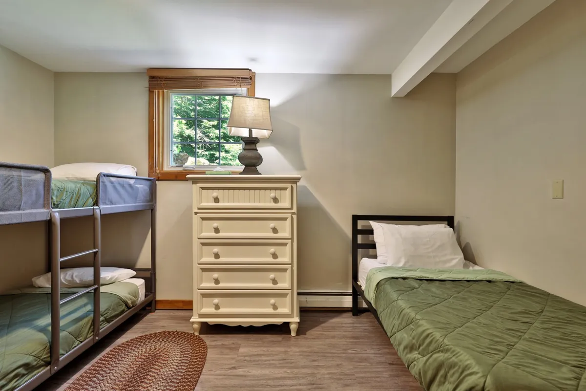 A cozy interior featuring bunk beds, a dresser, a lamp, and a bright window, creating a comfortable space for children. The arrangement suggests fun sleepovers, late-night chats, and a welcoming environment for kids to rest after adventurous days.