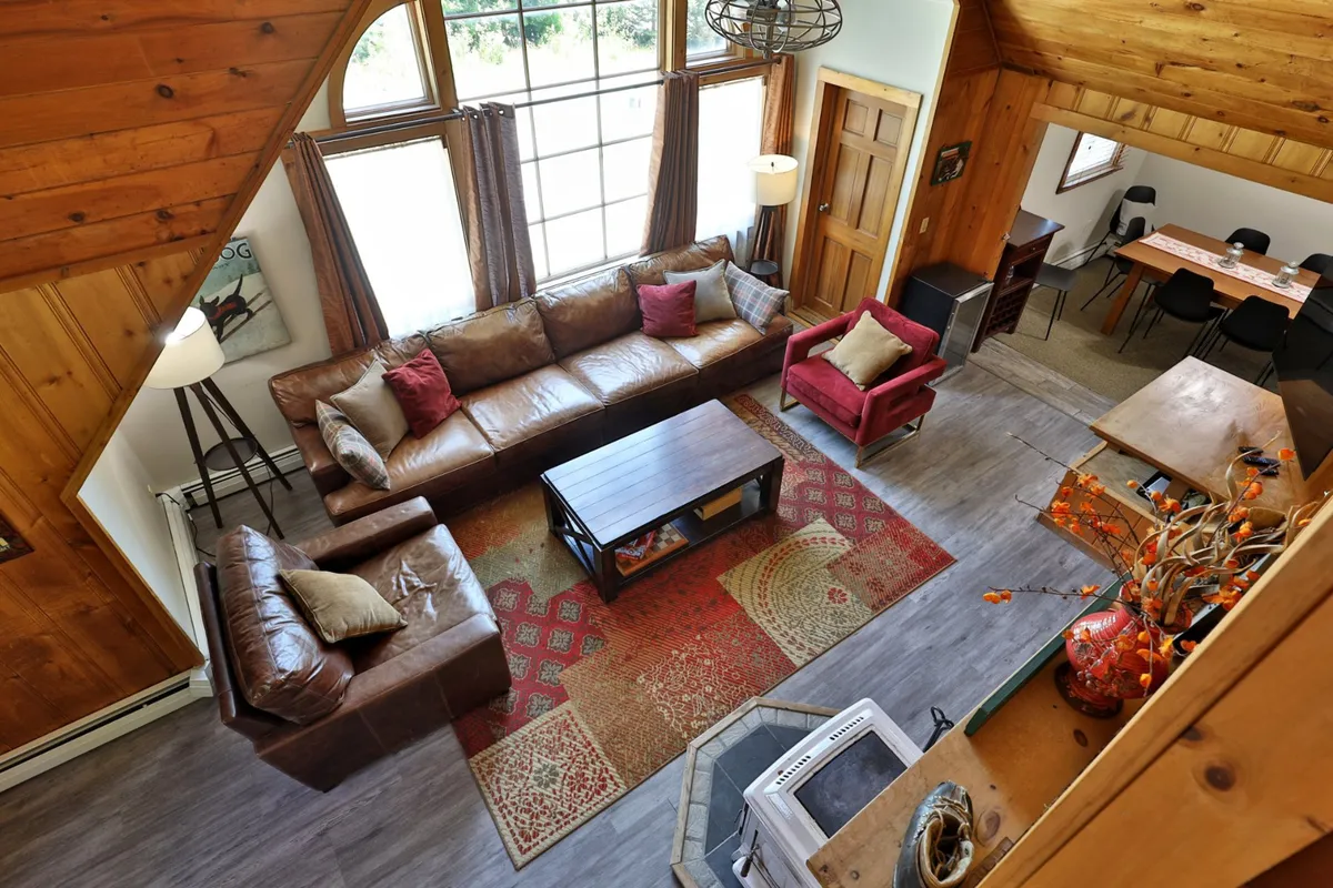 A spacious, light-filled living room with large windows that let in natural light, featuring comfortable leather sofas, colorful accent chairs, a stylish coffee table, and a decorative rug. The space is arranged for relaxation, entertainment, and group gatherings, with the smart TV subtly visible as part of the scene.