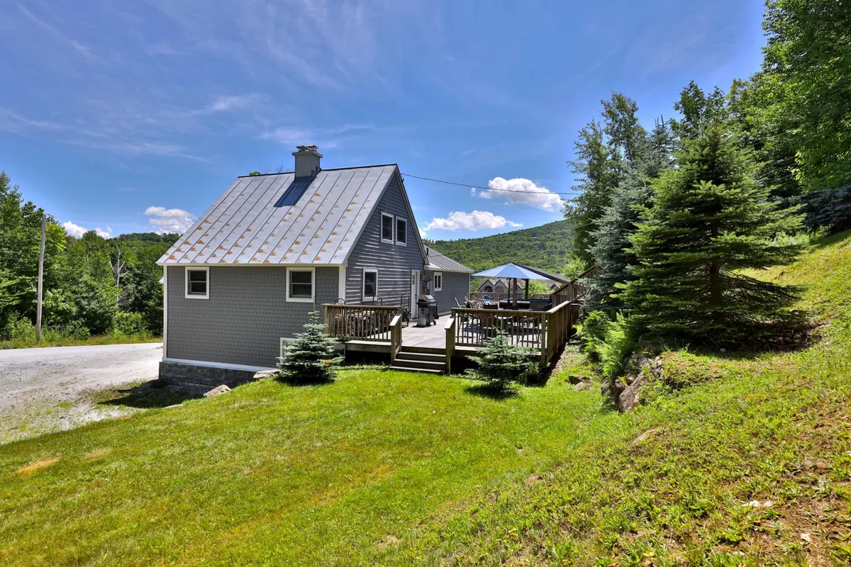 The chalet is nestled among tall trees, with a wooden deck overlooking the surrounding forest. The scene evokes peaceful outdoor living, with the deck suitable for morning coffee, sunsets, or watching pets roam safely while guests relax.