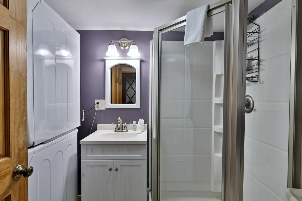 A well-organized bathroom with a vanity, sink, mirror, shower enclosure, and a stacked washer and dryer. The layout is practical, maximizing space for both bathing and laundry needs. The mood is efficient yet welcoming.